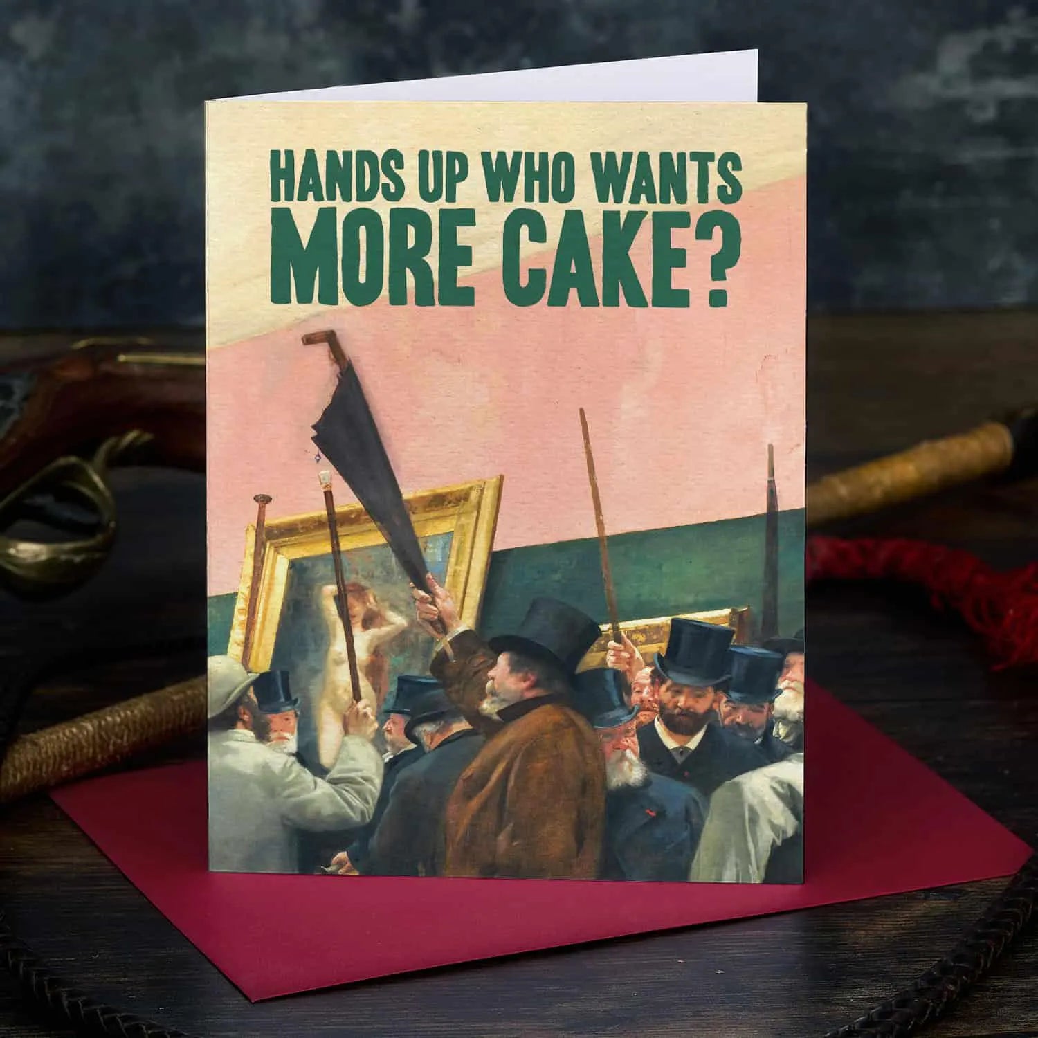 Card with vintage painting of people around a painting and text 'Hands up who wants more cake?' on a dark surface.