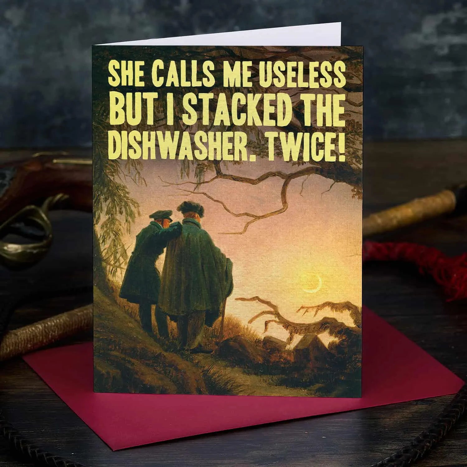 Greeting card with vintage-style illustration of two people embracing, text 'She calls me useless but I stacked the dishwasher twice!', and a dark background.