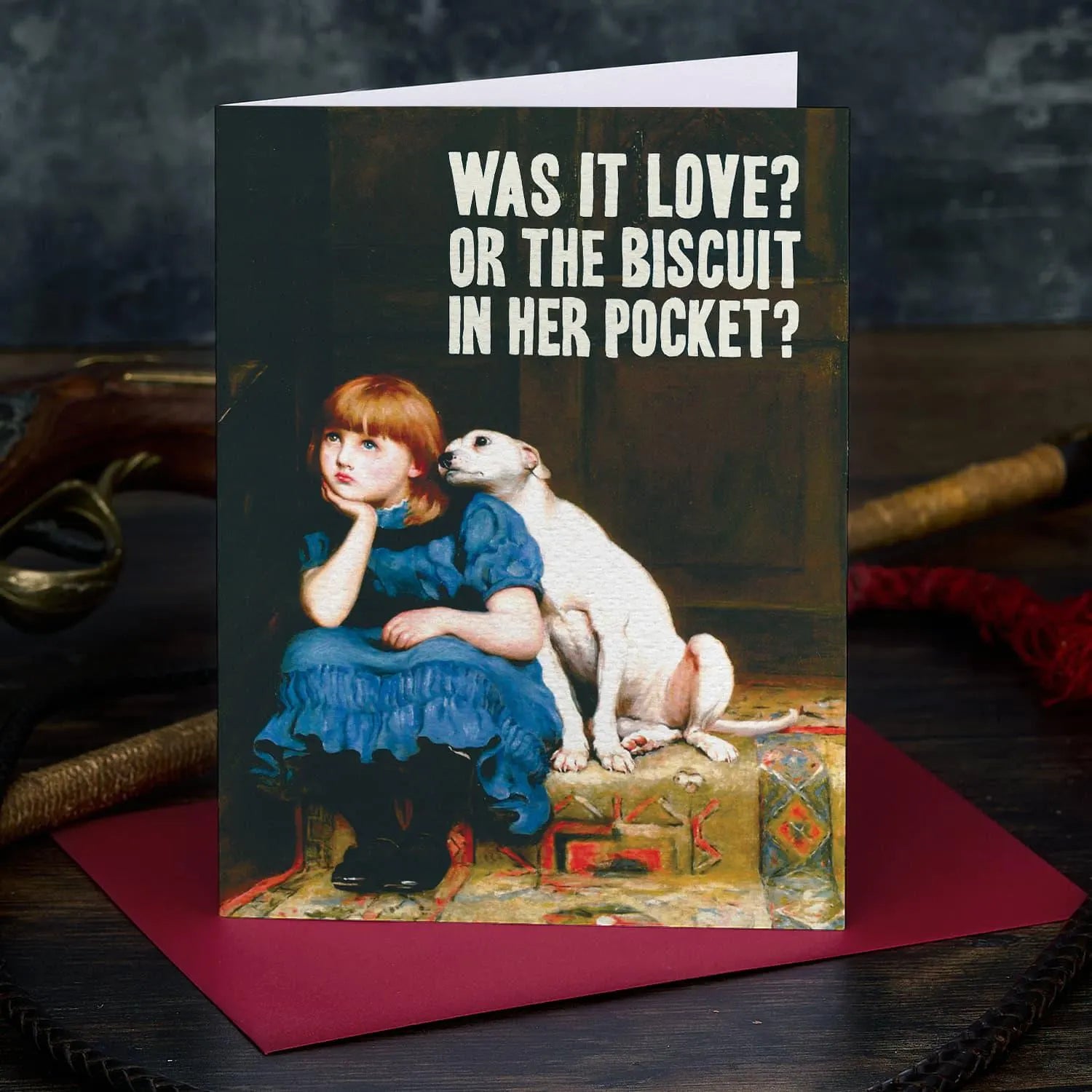Greeting card with a vintage-style illustration of a girl and dog, featuring text 'Was it love? Or the biscuit in her pocket?' on a dark background.