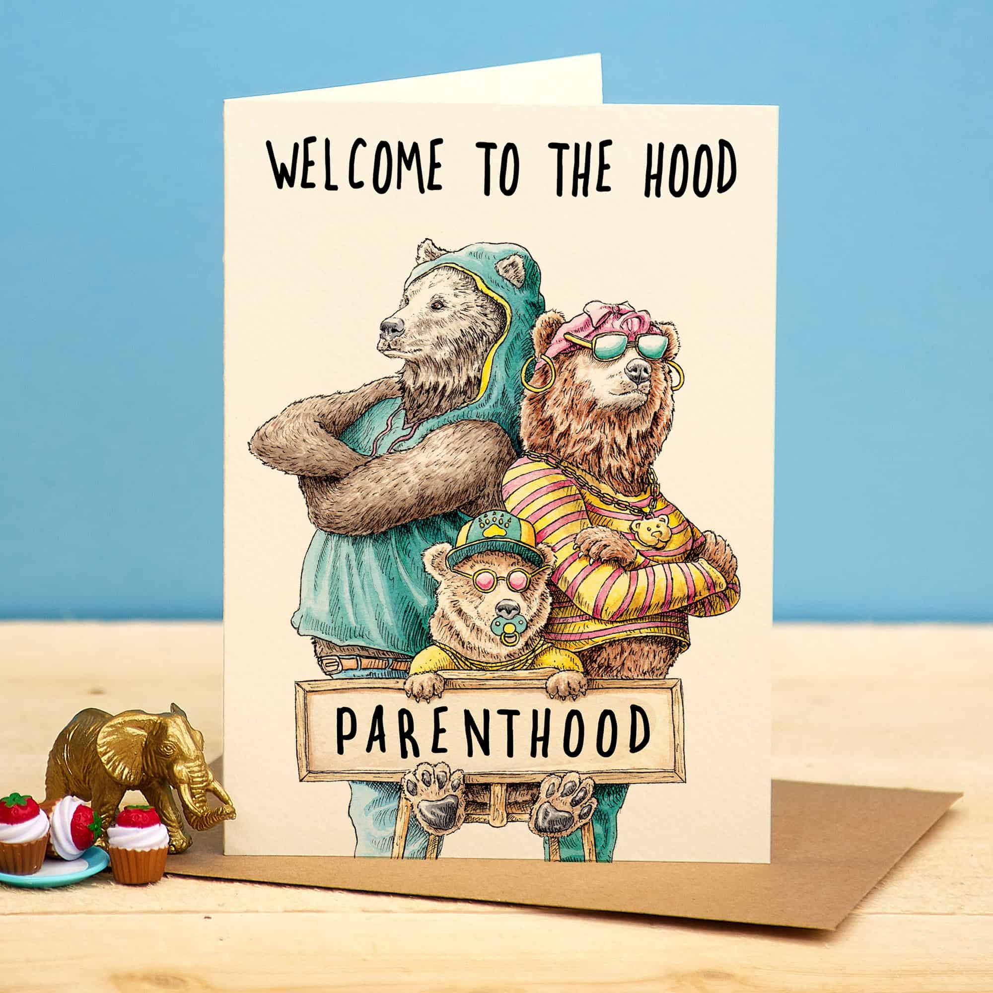 Card with cartoon bears holding a sign that reads 'Welcome to the hood Parenthood' on a wooden surface with a blue background.