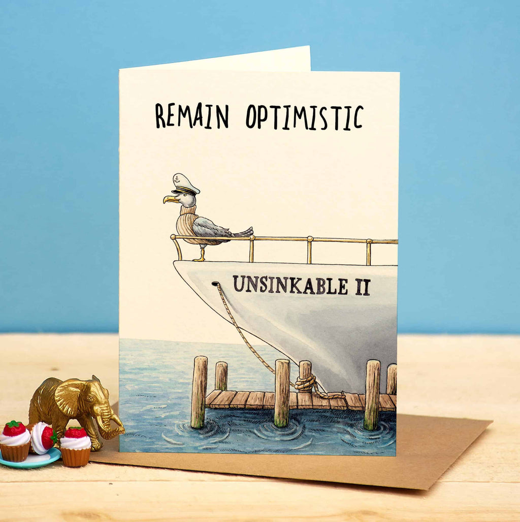 Card with a seagull on a ship labeled 'Unsinkable II' and text 'Remain Optimistic' on a blue and beige background.