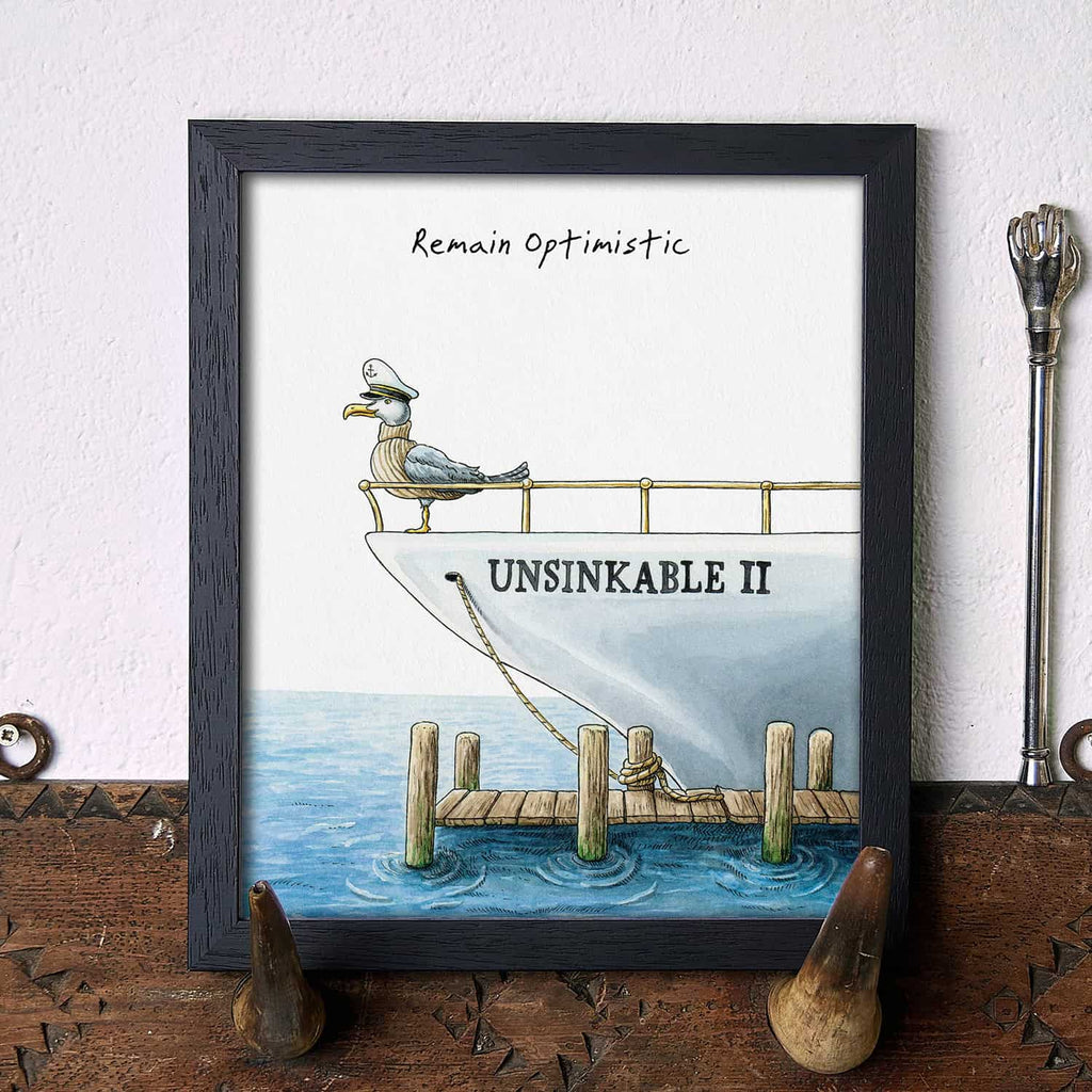 Artwork of a seagull on a boat with 'Unsinkable II' and 'Remain Optimistic' text, displayed on a wooden surface.