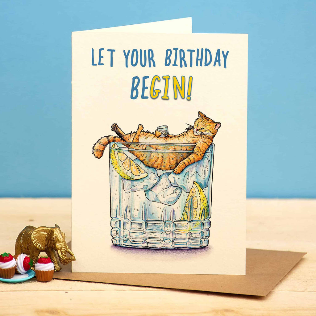 Birthday card with a cat in a glass of lemonade and ice, text 'Let your birthday begin!', on a blue background.