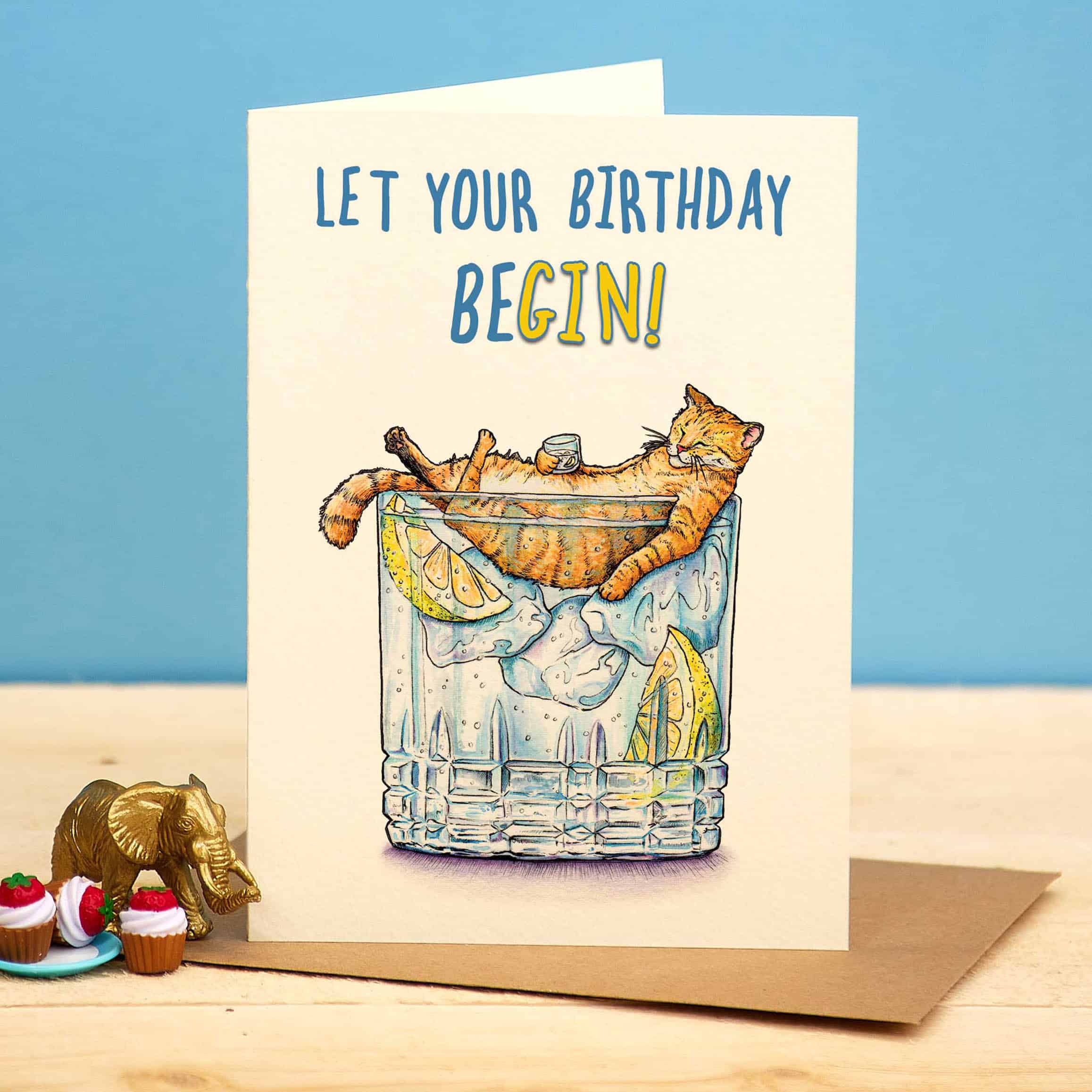 Birthday card with a cat in a glass of lemonade and ice, text 'Let your birthday begin!', on a blue background.