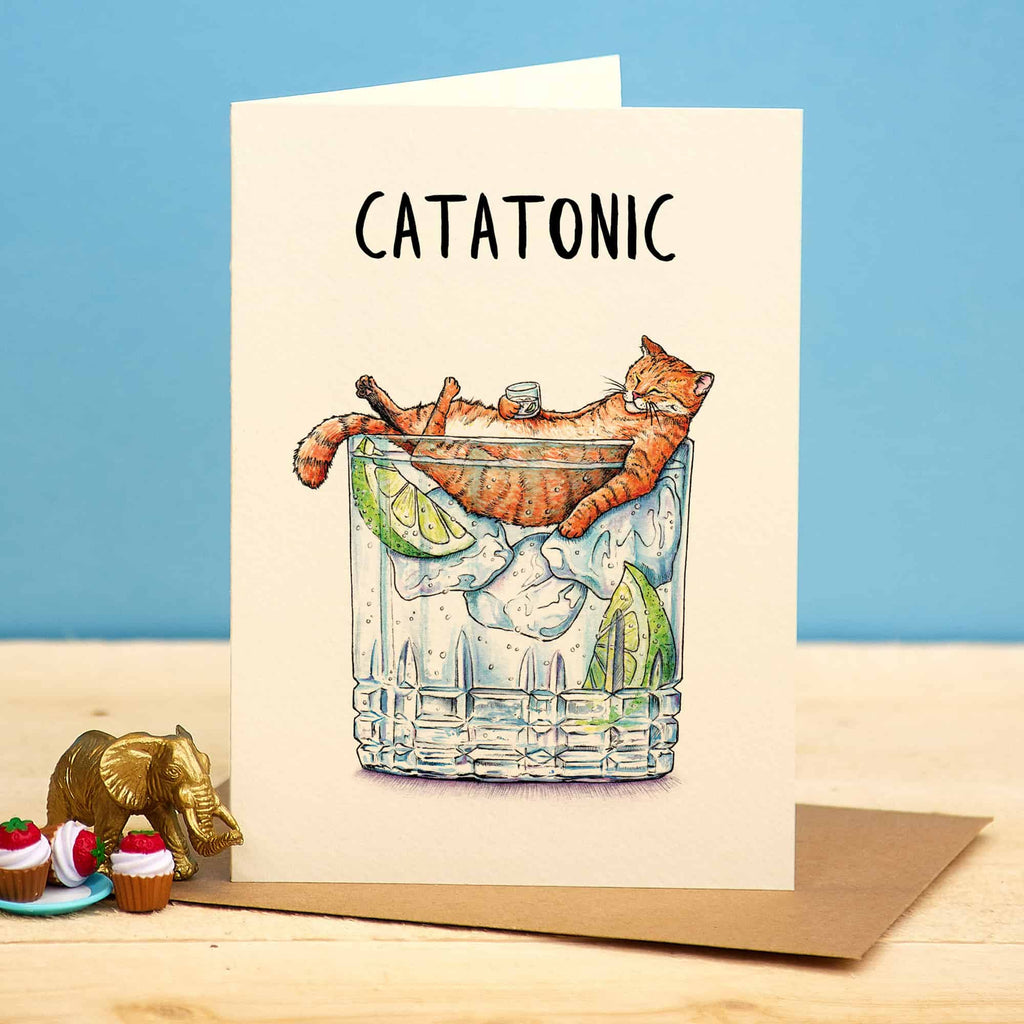 Card with a cat in a glass of gin and tonic, labeled 'Catatonic', on a wooden surface with a blue background.