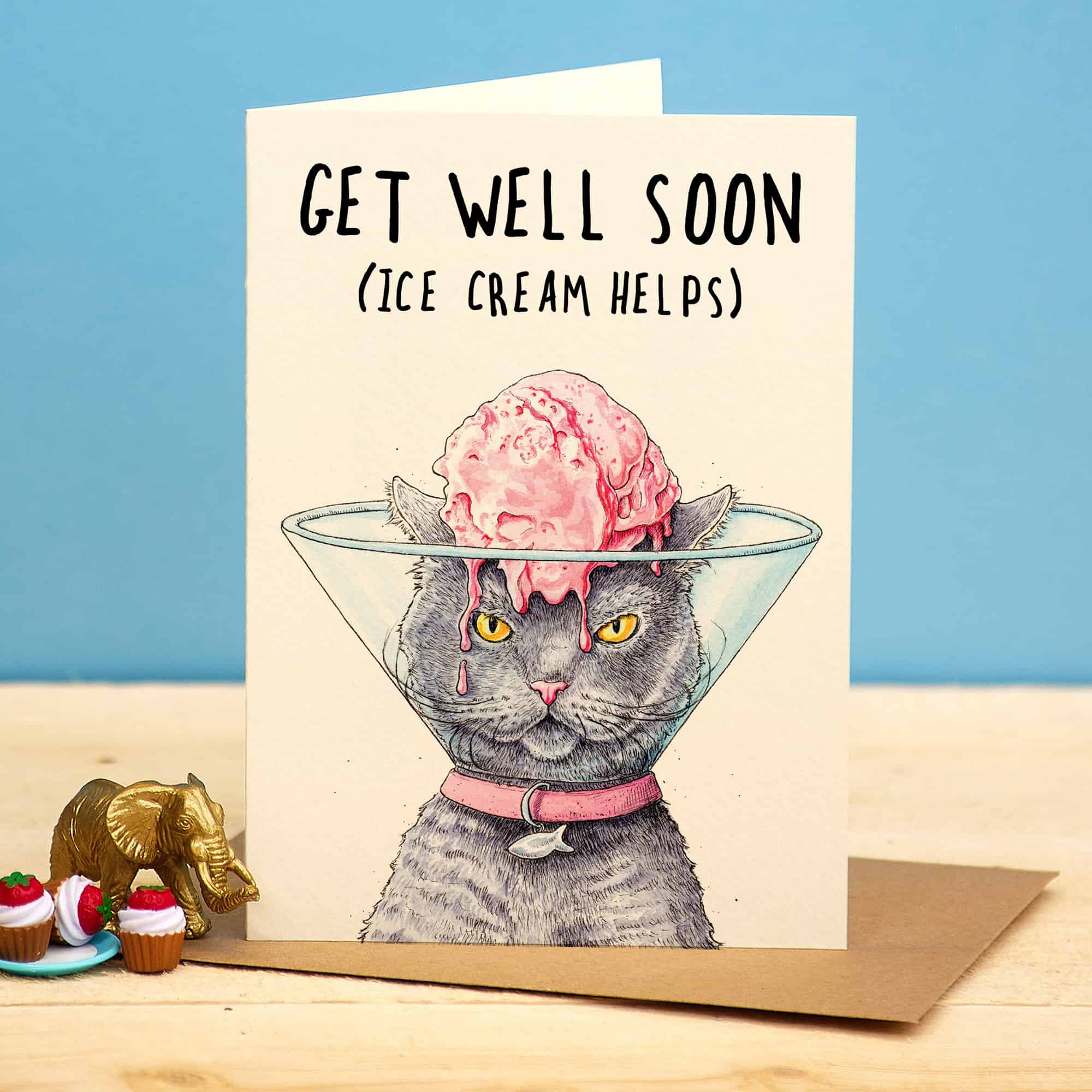 Get well soon card with a cat wearing a cone and ice cream on its head, on a wooden surface with a blue background.