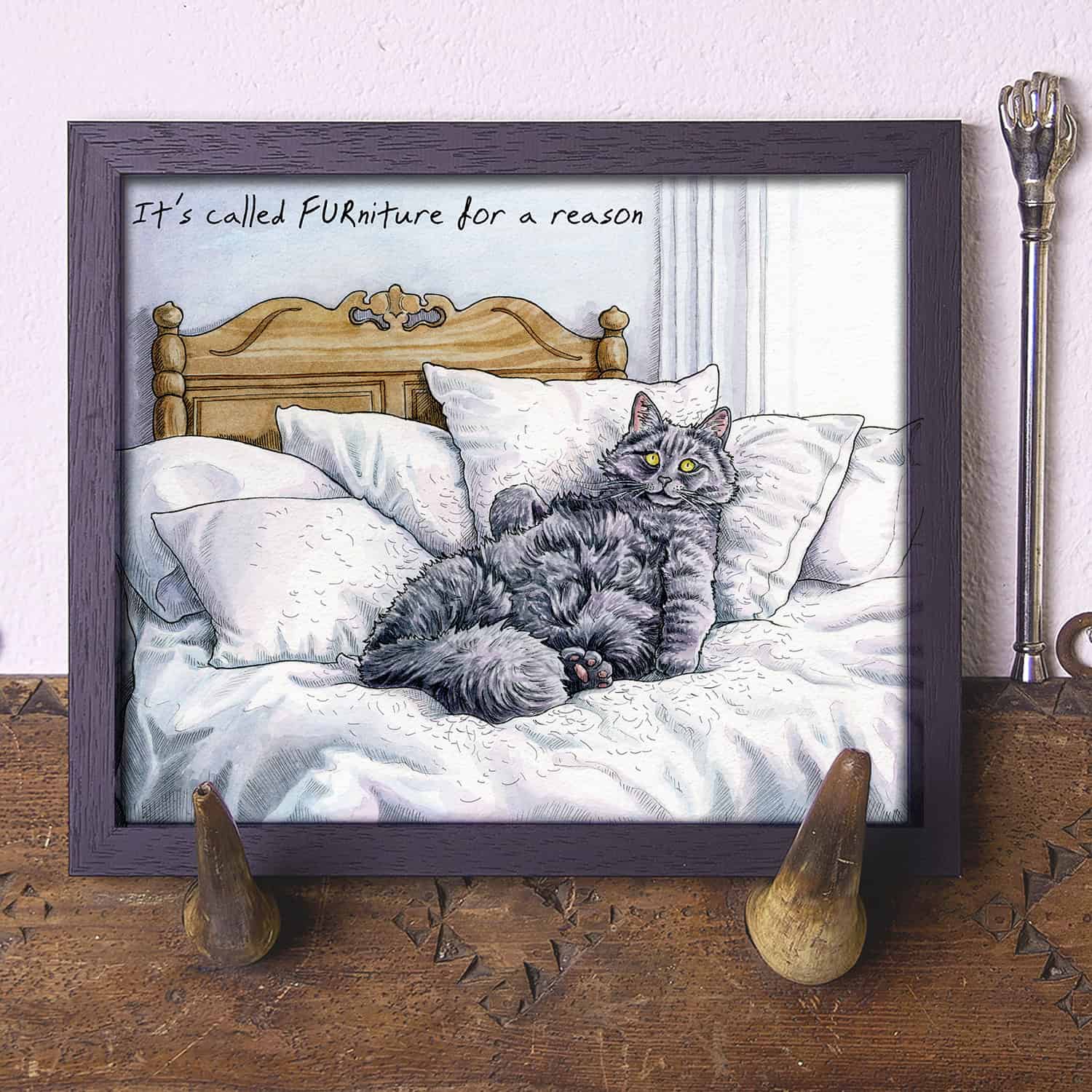 Artwork of a cat lying on furniture with humorous text, displayed on a wooden surface.