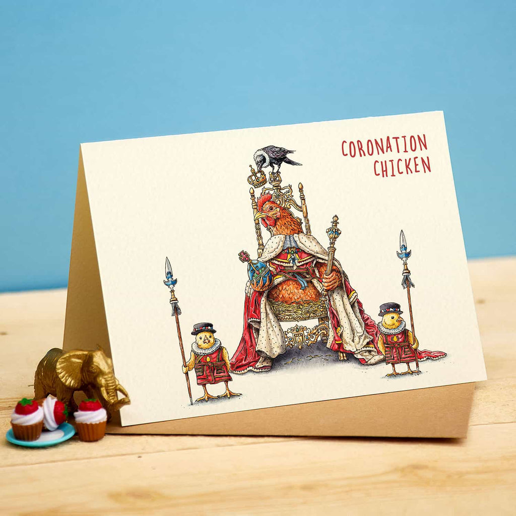 Card with a chicken on a throne labeled 'Coronation Chicken' on a wooden surface with a blue background.