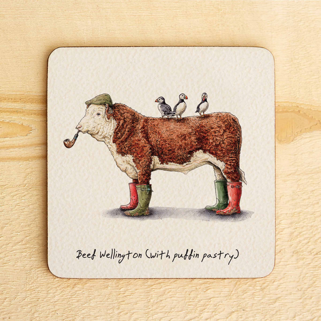 Coaster with illustration of a cow in Wellington boots with puffins on its back, on a wooden surface.