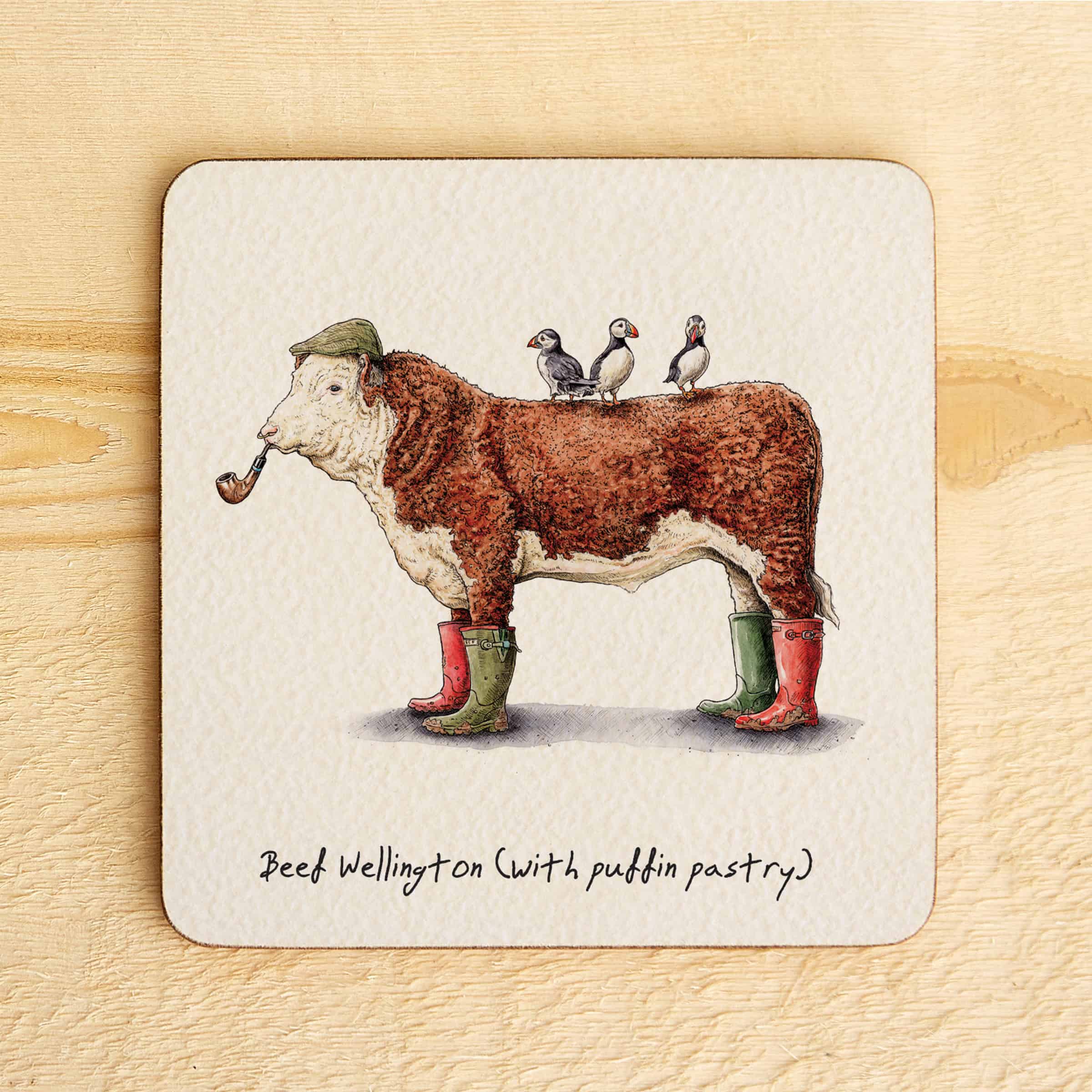 Coaster with illustration of a cow in Wellington boots with puffins on its back, on a wooden surface.