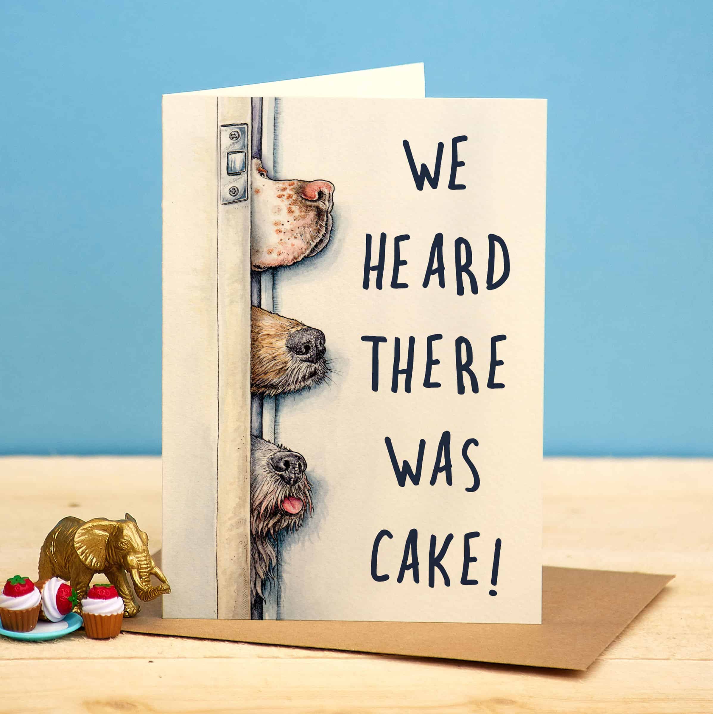 Greeting card with dogs peeking around a door and text 'We heard there was cake!' on a blue background.
