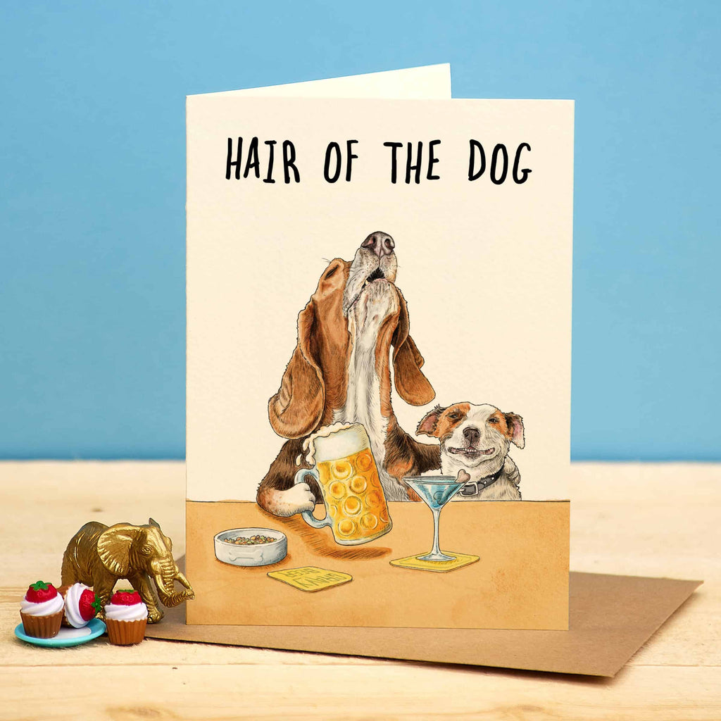 Card with illustration of two dogs and a drink, with text 'Hair of the Dog', on a wooden surface with a blue background.