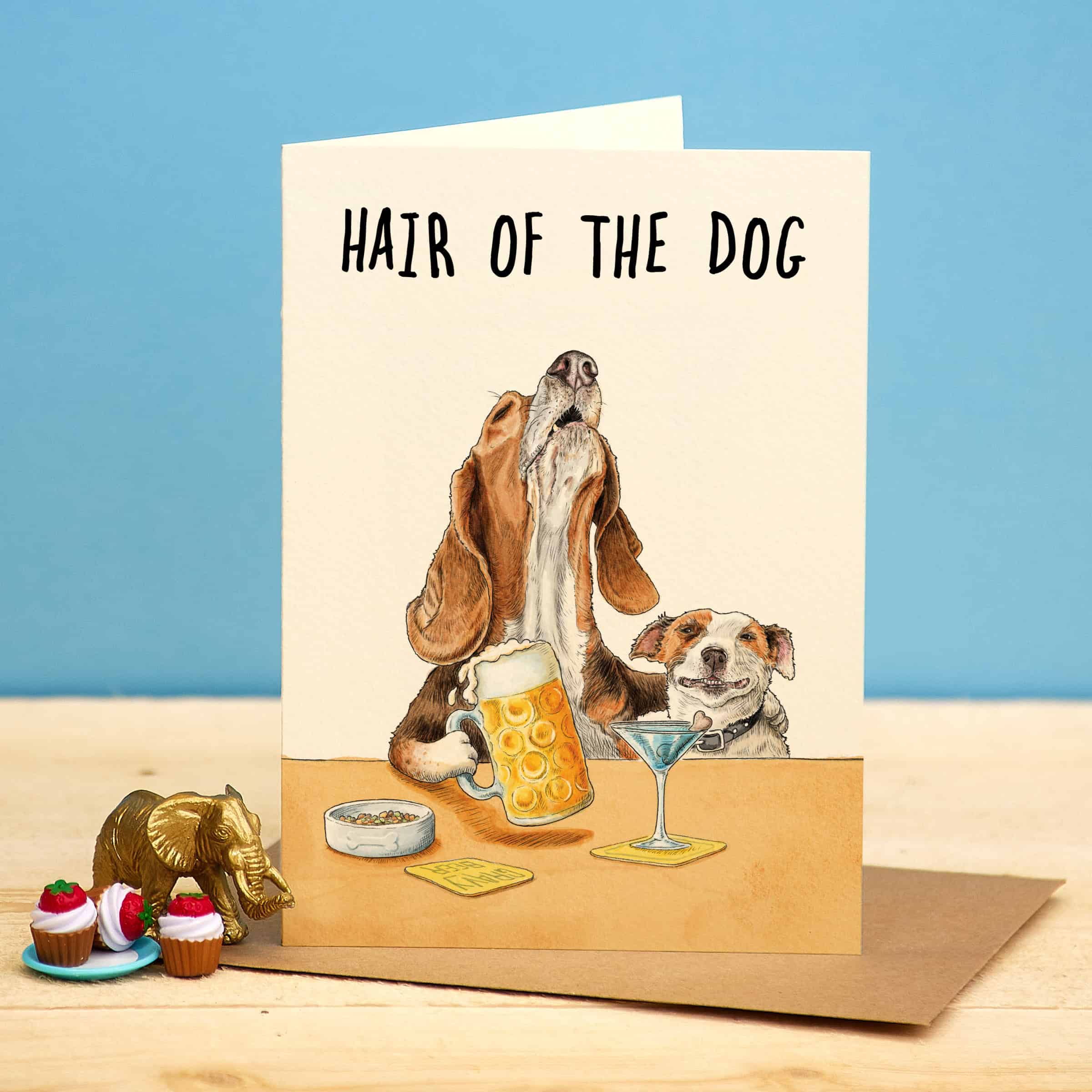 Card with illustration of two dogs and a drink, with text 'Hair of the Dog', on a wooden surface with a blue background.