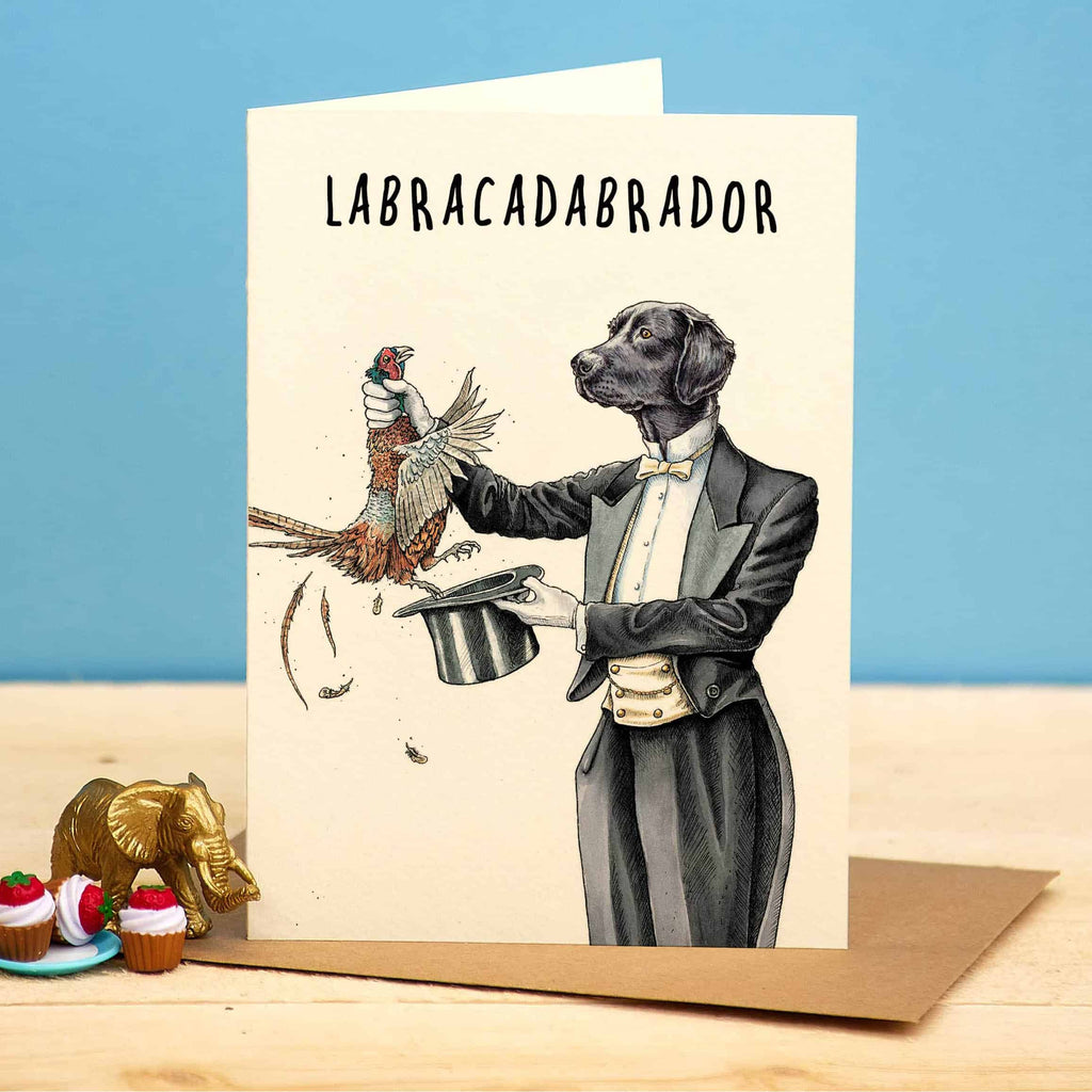 Card with a dog in a tuxedo holding a pheasant and top hat, with 'LABRACADABRADOR' text, on a wooden surface with decorative items.