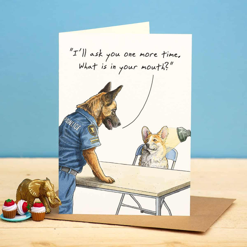 Greeting card with illustration of a police dog and a corgi at a table, with text 'I'll ask you one more time. What is in your mouth?' on a blue background.