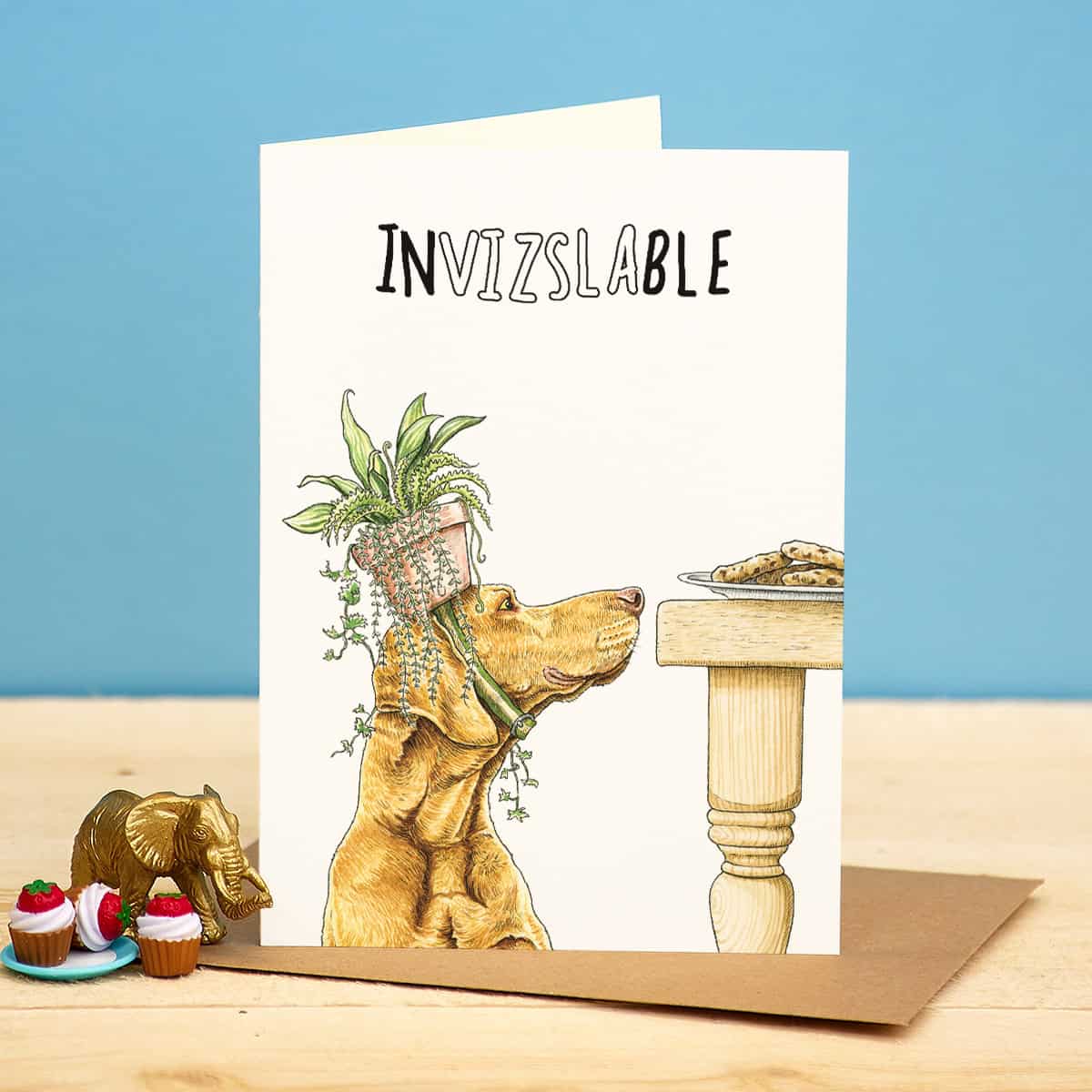 Card with a dog holding a plant, titled 'INVIZSLABLE', on a wooden surface with a blue background.