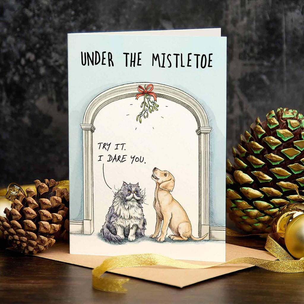 Christmas card with two dogs under a mistletoe arch, surrounded by pine cones and a gold bow.