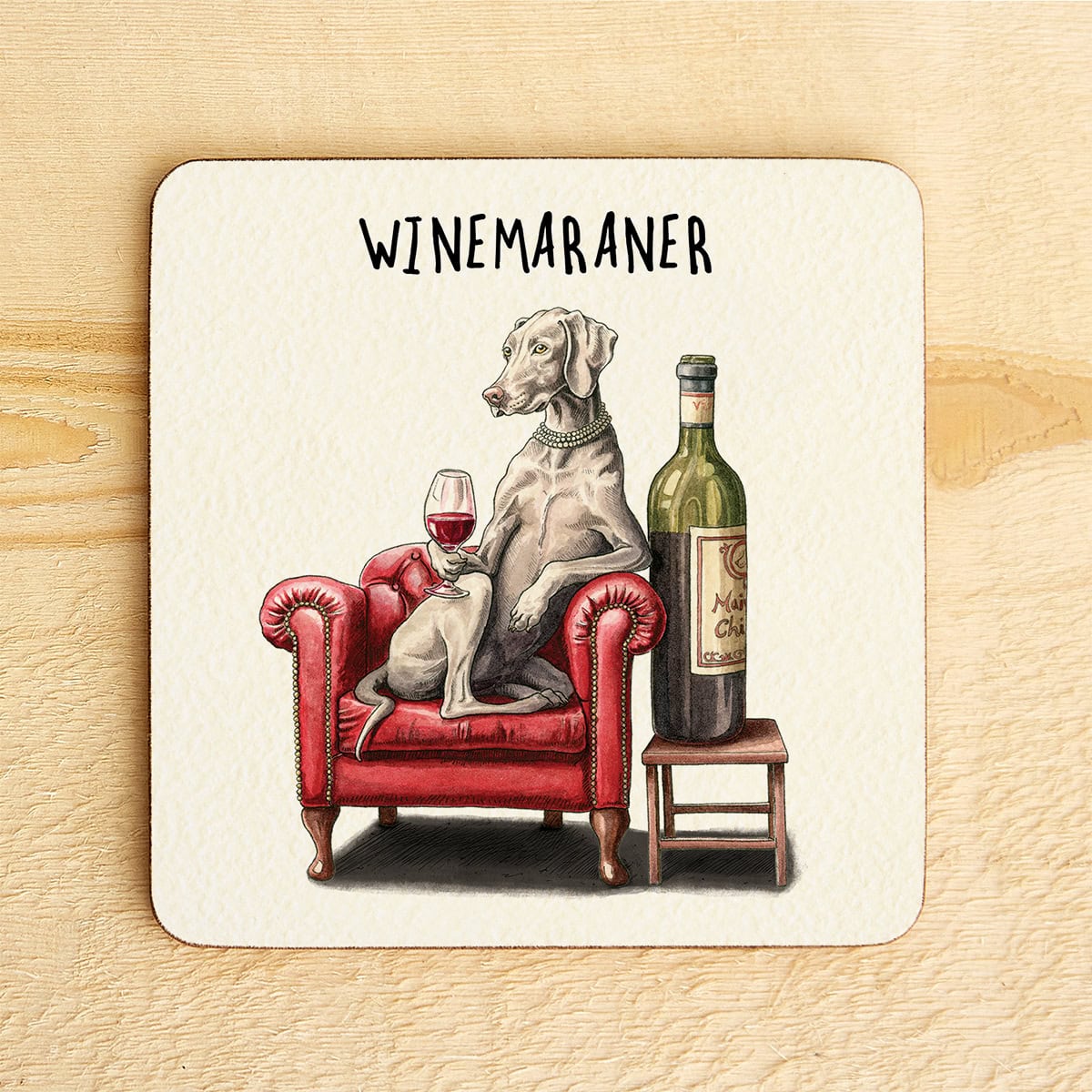 Coaster with a Weimaraner dog sitting on a red chair holding a wine glass, next to a bottle of wine, on a wooden surface.