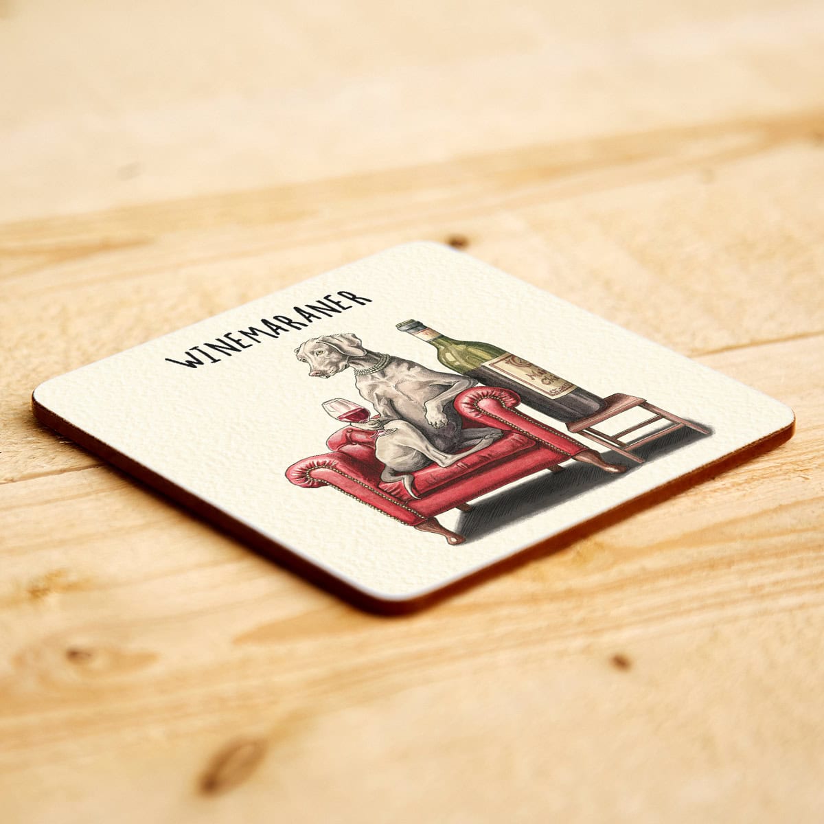 Coaster with a graphic design of a Weimaraner dog sitting on a chair with a bottle, placed on a wooden surface.