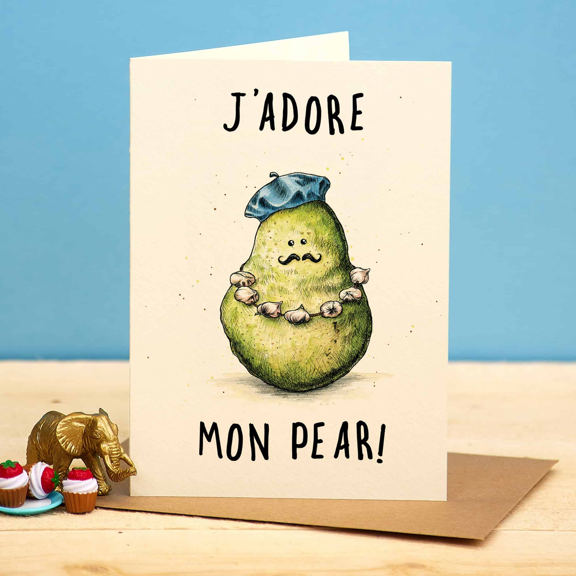 Card with a pear illustration wearing a beret and necklace, text 'J'ADORE MON PEAR!', on a wooden surface with decorative items.