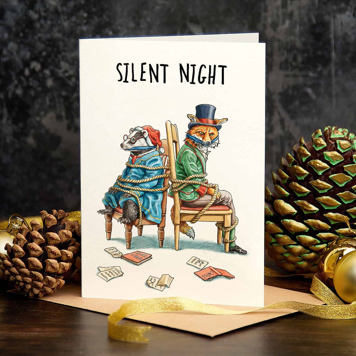 Greeting card with illustration of animals in costumes and 'Silent Night' text, surrounded by pine cones and a gold ribbon.