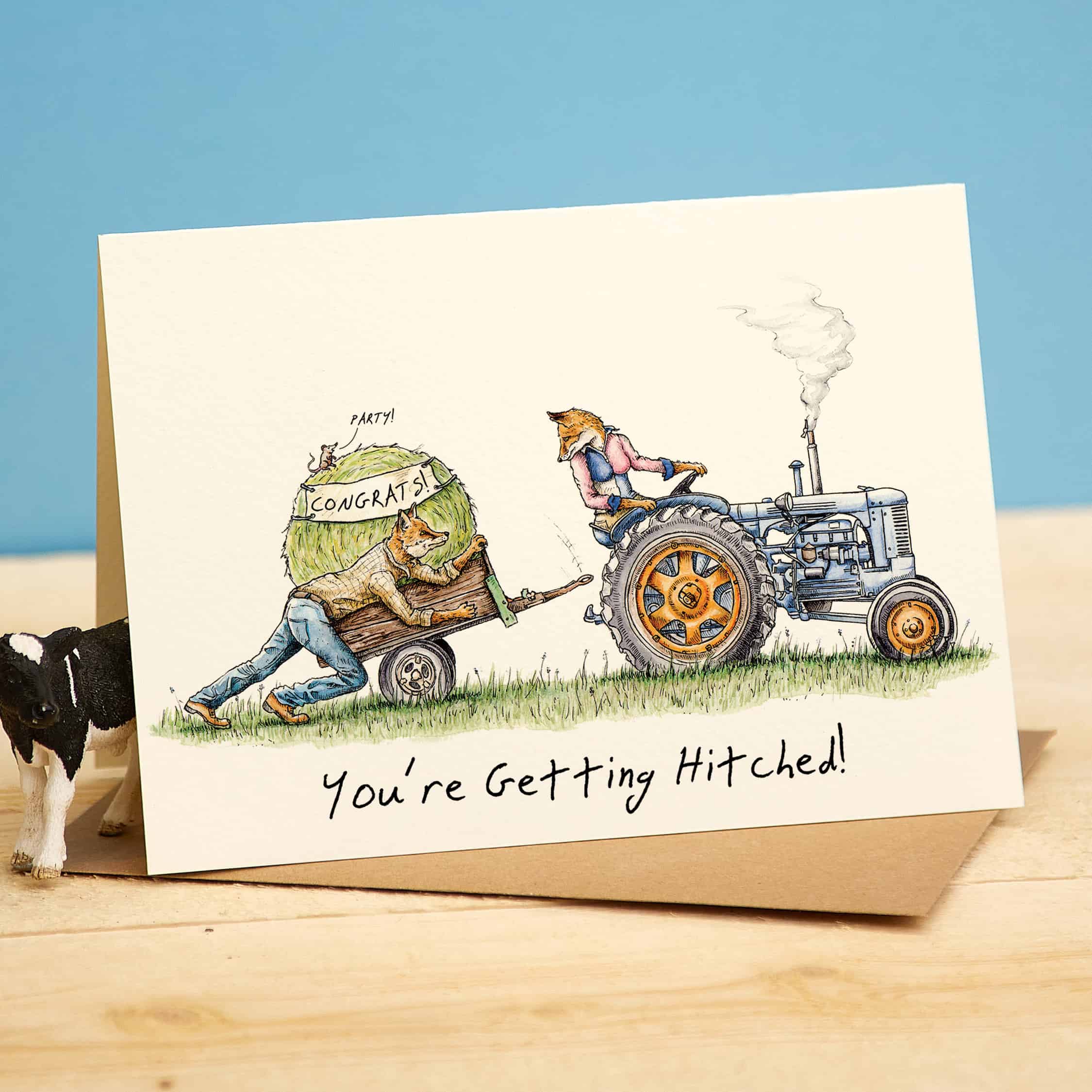 Card with illustration of a fox on a tractor and fox in a cart, with text 'You're Getting Hitched!' on a wooden surface.