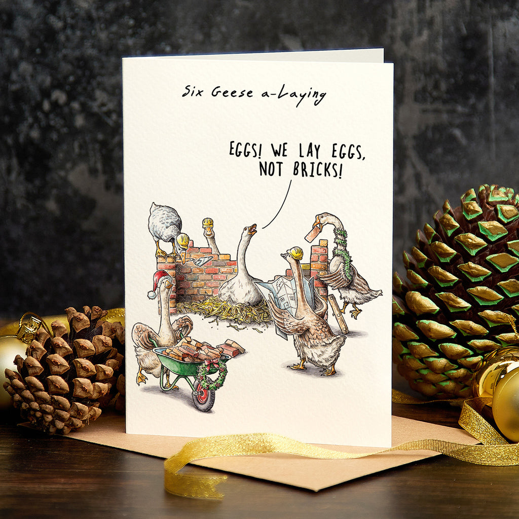 Card with illustration of geese and humorous text, surrounded by pine cones and a gold ribbon on a dark background.