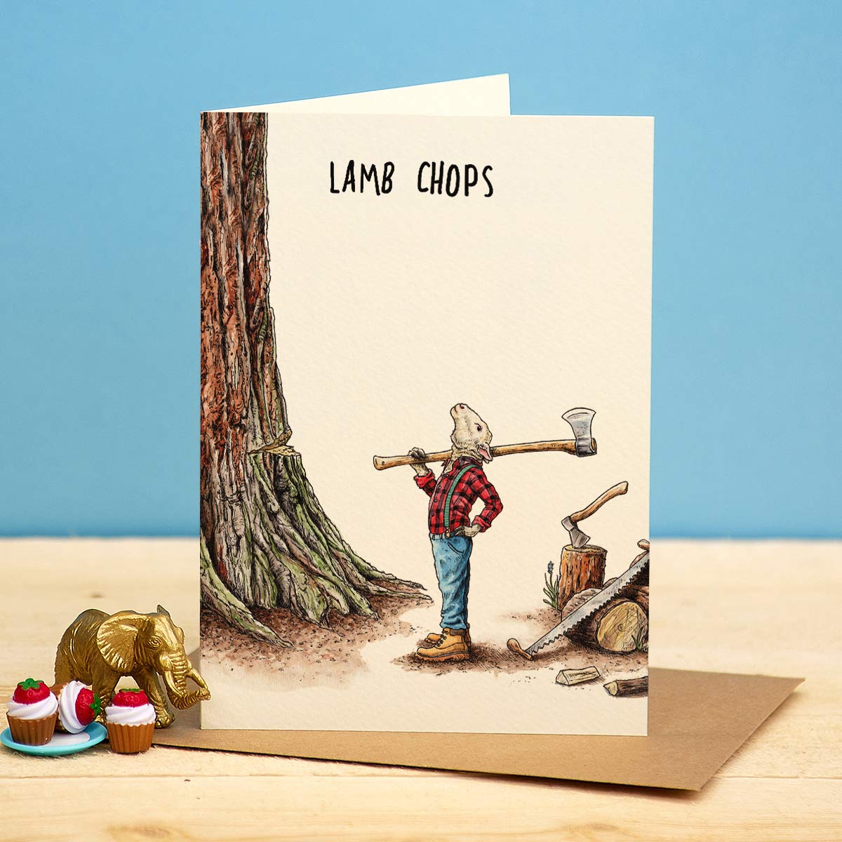 Greeting card with a bear holding a log over its shoulder, labeled 'Lamb Chops', on a wooden surface with a blue background.