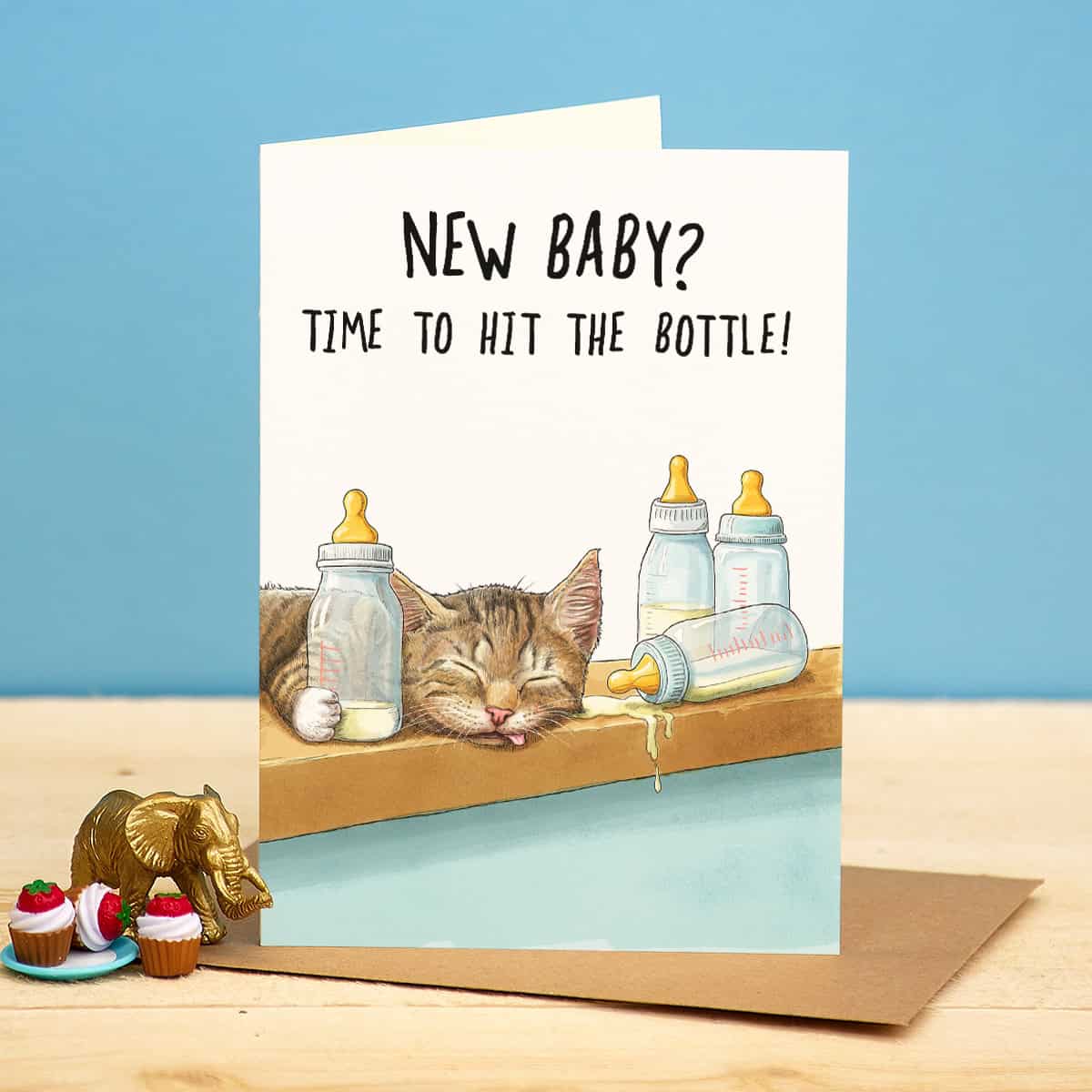 Card with a cat and baby bottles on a wooden surface, text 'NEW BABY? TIME TO HIT THE BOTTLE!' against a blue background.