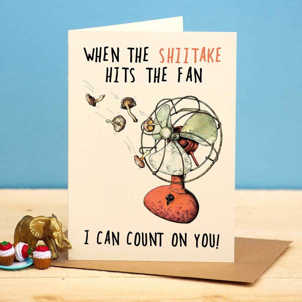 Greeting card with a fan illustration and humorous text on a wooden surface with a blue background