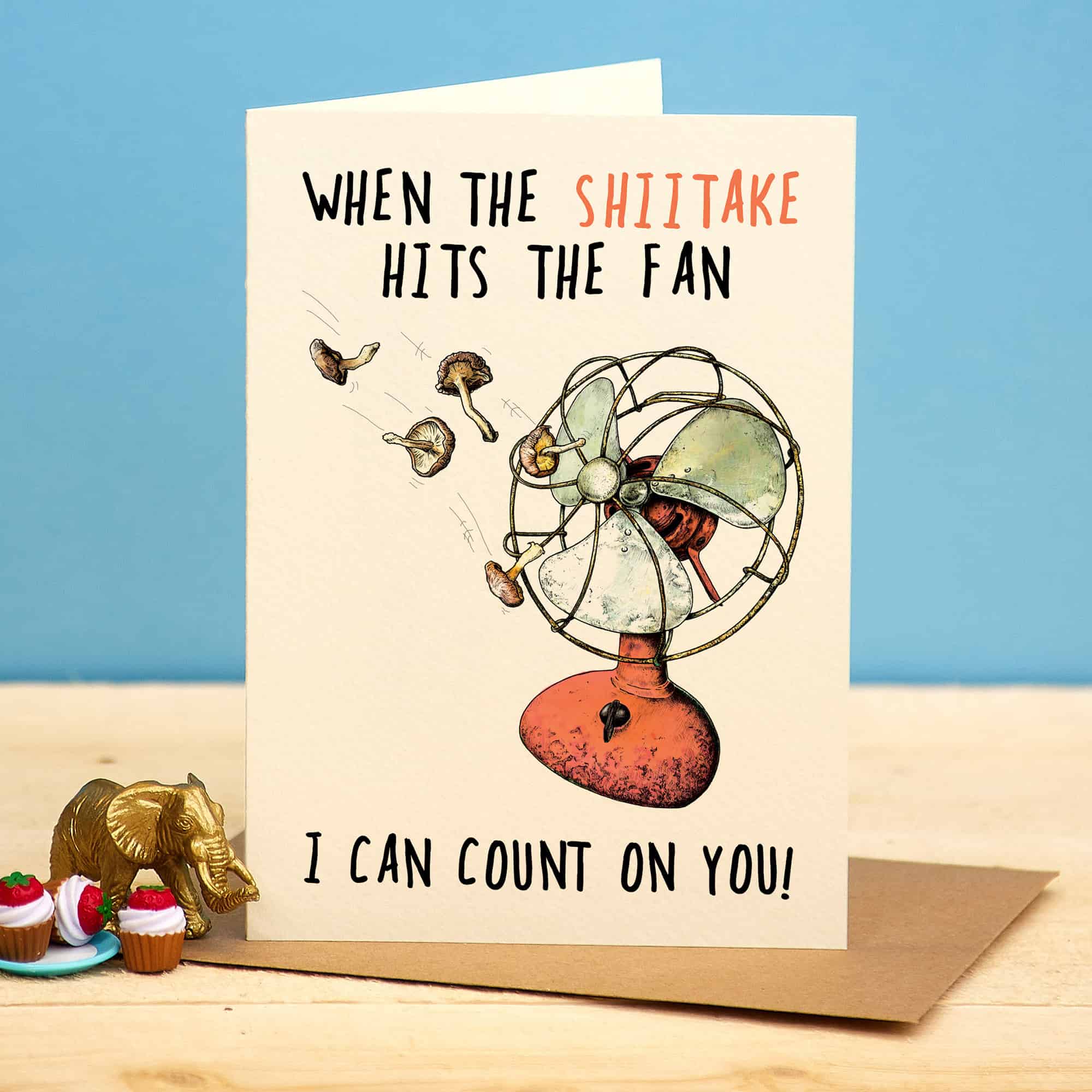 Greeting card with a fan illustration and humorous text on a wooden surface with a blue background