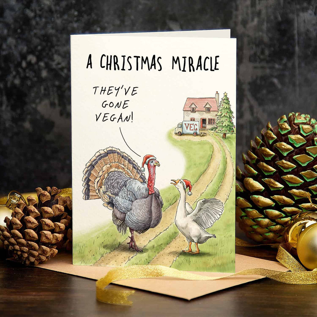 Card with cartoon turkeys and goose on a road, text 'A Christmas Miracle They've Gone Vegan', surrounded by pine cones and gold ornaments.