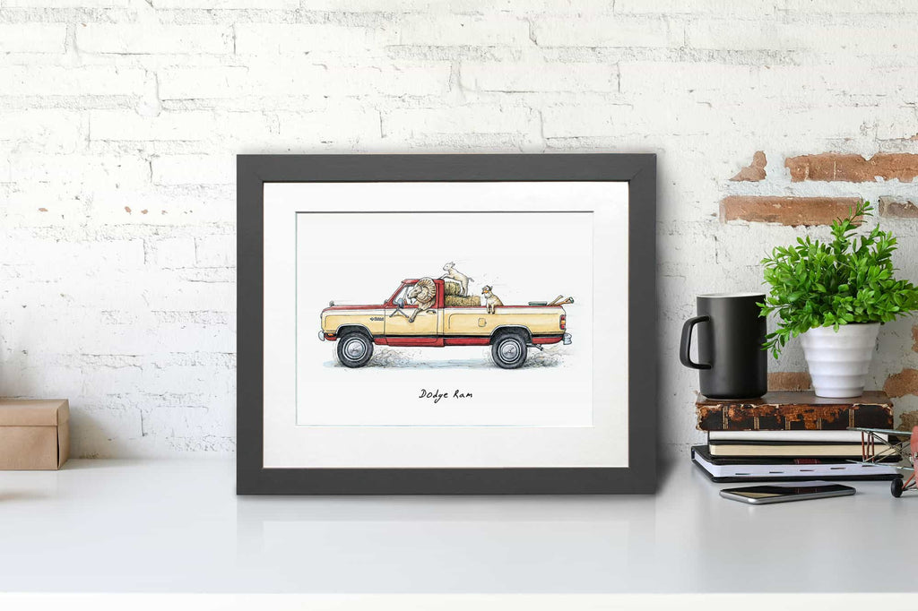 Print of a Dodge Ram pickup truck being driven by a ram with two lambs in the back in a grey frame sitting on a white desk
