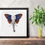 Print of an elephant with colourful butterfly wings in a black frame on a wooden shelf leaning against a wall