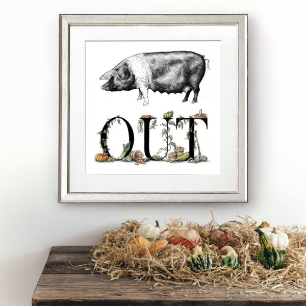 Print of a saddleback pig and lettering decorated with vegetables in a white frame on a white wall above a wooden shelf