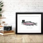 Print of a fish wearing a top hat with a monocle in a black frame on a wooden desk leaning against a white brick wall