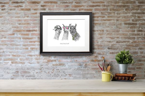 Print of three black and white llamas wearing sunglasses, a cap and a gold chain in a black frame on a red brick wall