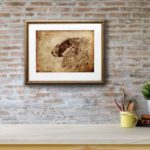 Print of a sheep’s head on an aged textured background in a gold frame on a red brick wall above a white shelf