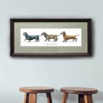 Print of three dachshunds holding each other’s tails in a line in a dark wood frame on a wall above stalls