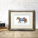 Original artwork of a Shetland pony in a blue dress in a silver frame resting on a wooden shelf against a white brick wall